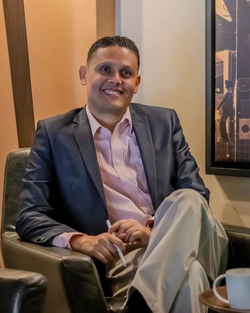 Rolando López, sentado y sonriendo, con chaqueta oscura y camisa rosada.